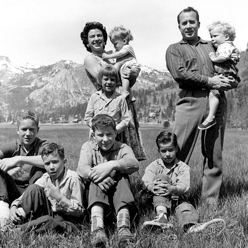 A group of nine people, including adults and children, pose outdoors with mountains in the background, sitting and standing on a grassy field.