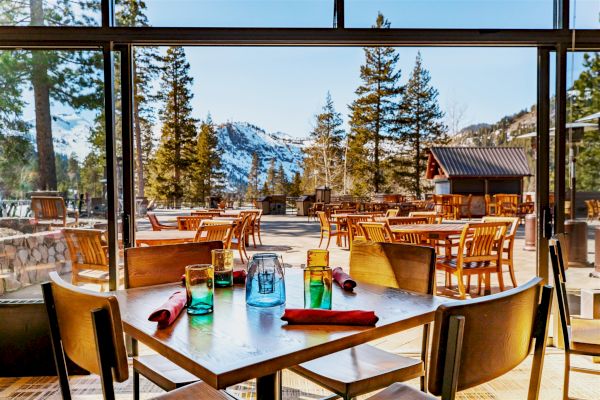 The image shows a dining area in a restaurant with wooden tables and chairs, set against a stunning mountain and forest backdrop with snow.