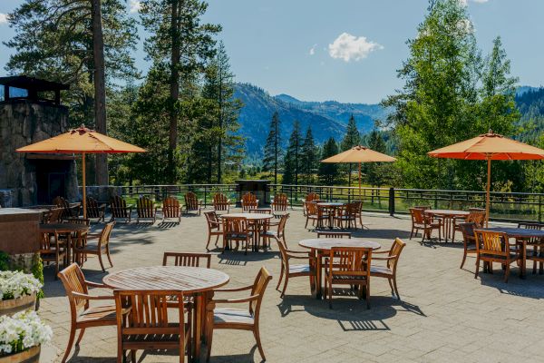 An outdoor patio with tables, chairs, and large umbrellas overlooks a scenic mountain view surrounded by tall trees and a clear sky.
