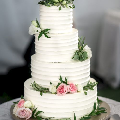 A three-tiered white cake decorated with pink and white flowers and green leaves stands on a table with plates and cutlery.