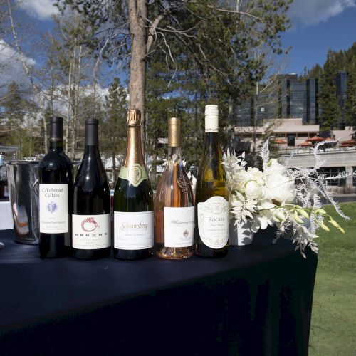 The image shows a table outdoors with six wine bottles, white flowers, and a sunny background with trees and buildings.