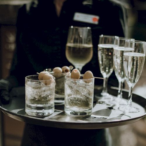 A server holds a tray with drinks: three glasses of sparkling wine, one glass of white wine, and two cocktails garnished with olives.