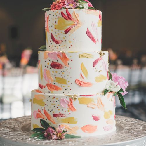 This image features a three-tiered wedding cake adorned with colorful brushstroke patterns and topped with flowers, placed on an ornate stand.