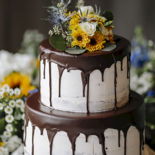 A two-tier cake with white frosting and chocolate drip, topped with a flower arrangement of yellow, blue, and white blooms, on a laced cake stand.