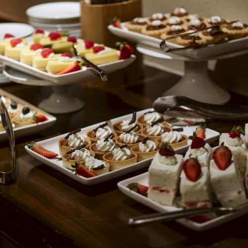 A buffet table with various desserts, including cakes, tarts, and pastries, each garnished with fruits like strawberries and blueberries.