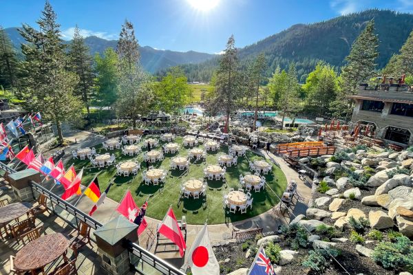 The image shows an outdoor event setup with multiple round tables and international flags, set amidst a scenic mountain landscape with evergreen trees and rocks.