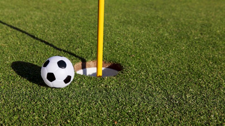 A soccer ball is positioned next to a hole on a golf course, with a flagpole in the hole, blending elements of both sports perfectly.