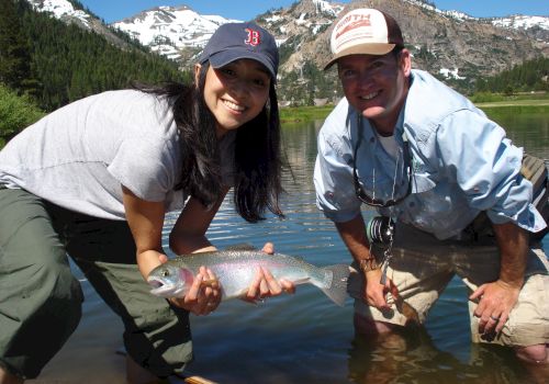 Two people are standing in a lake with a mountainous background, holding a freshly caught fish, and smiling for the camera.