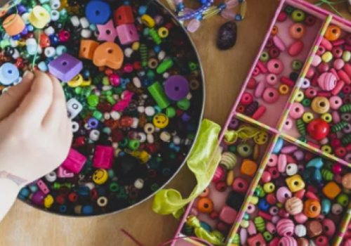A hand is selecting beads from a bowl filled with colorful beads, with a pink organizer containing more sorted beads and materials on the side.
