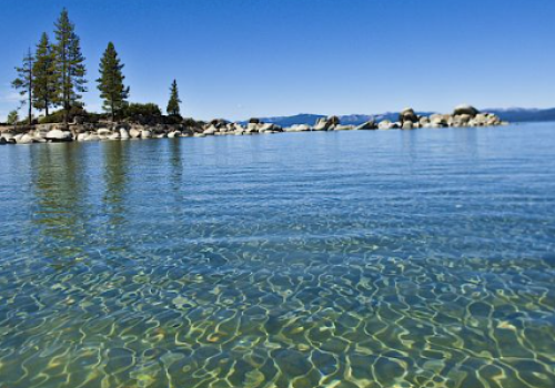The image shows a serene lake with clear water, rocky shoreline, and trees in the background under a clear blue sky.